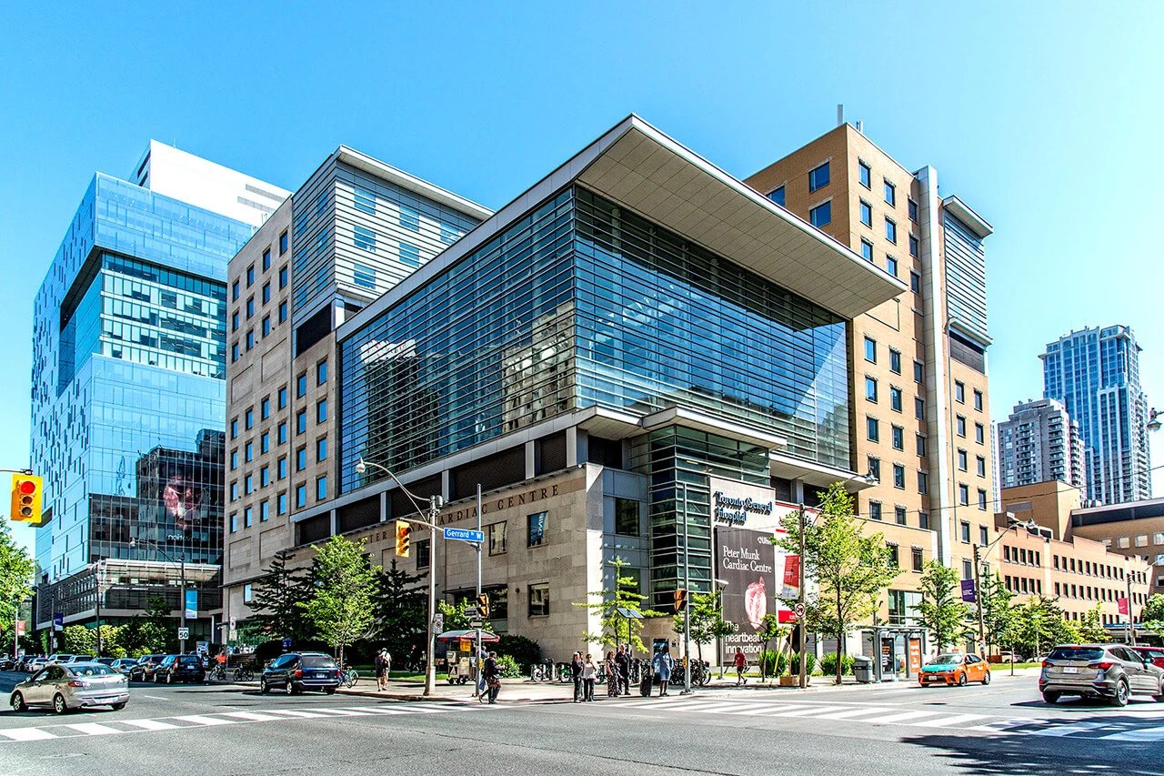 Modern cityscape showing a large glass-fronted building with a unique overhanging architectural design on a sunny day, surrounded by bustling street traffic and pedestrians, prominently featuring the UHN Foundation headquarters.